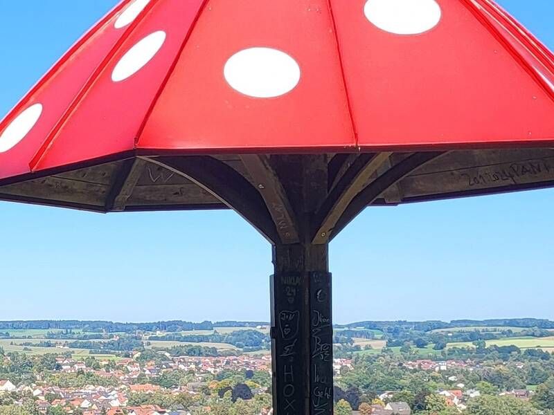 Ein großer, roter Pavillon in Form eines Fliegenpilz-Hutes mit weißen Punkten steht auf einer Anhöhe mit Blick auf eine ländliche Landschaft.