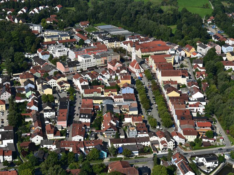 Eine Luftaufnahme zeigt eine malerische Stadt mit dicht aneinander gebauten Häusern und roten Dächern, umgeben von grünen Bäumen und Wiesen.