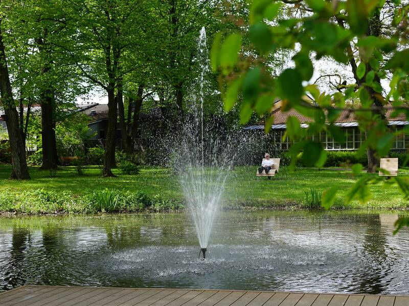 Ein Springbrunnen in einem Teich sprudelt in einem grünen Park, während eine Person auf einer Bank im Hintergrund sitzt.