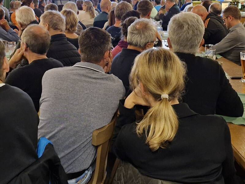 Eine Gruppe von Menschen sitzt dicht gedrängt an Tischen in einem Raum und hört einer Versammlung zu, wobei Biergläser auf den Tischen stehen.