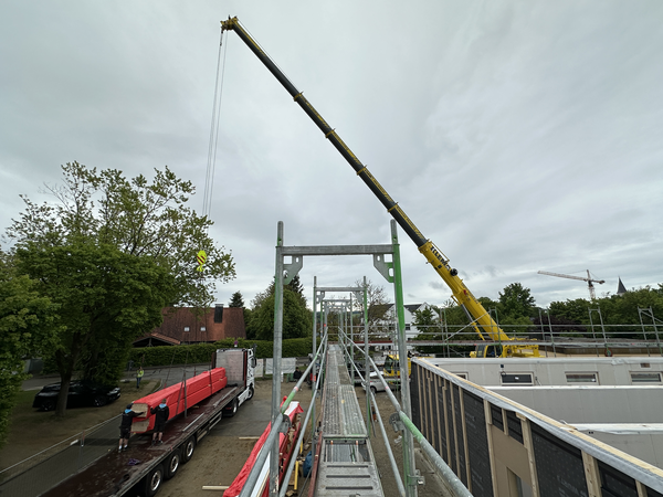 Ein Kran hebt Teile von einem Transporter. Rechts im Bild ist der Neubau auf den die Balken gehoben werden.