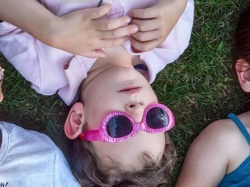 Drei Kinder liegen mit herzförmigen Sonnenbrillen auf einer Wiese und schauen in den Himmel.