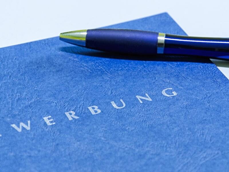 Ein blauer Ordner mit der Aufschrift "BEWERBUNG" liegt auf einem Tisch und ein blauer Kugelschreiber darauf.