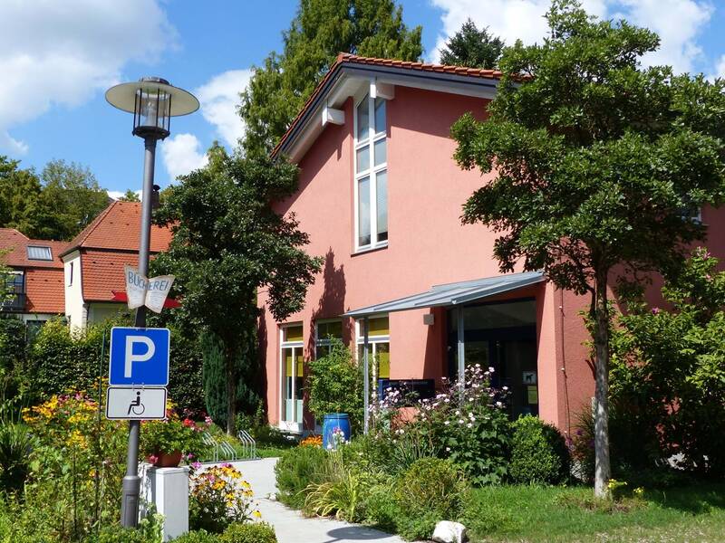 Im Bild ist ein Gebäudeeingang zu einem rosa Haus mit einem Vordach zu sehen. Es führt ein gepflasteter Weg zum Haus gesäumt von einem Garten mit Sträuchern und bunten Blumen.