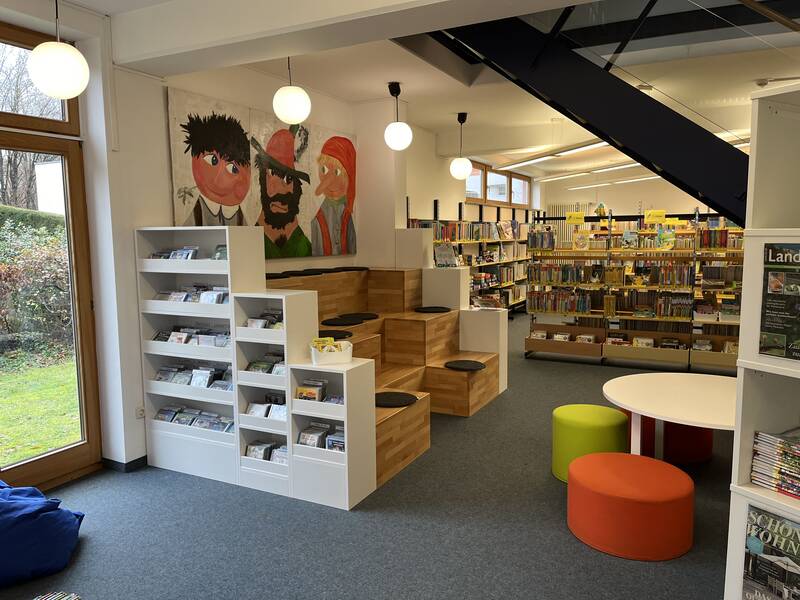Eine gemütliche und bunte Bibliotheksecke mit Regalen voller Bücher, Sitzgelegenheiten und großen Fenstern, durch die man ins Grüne blickt.