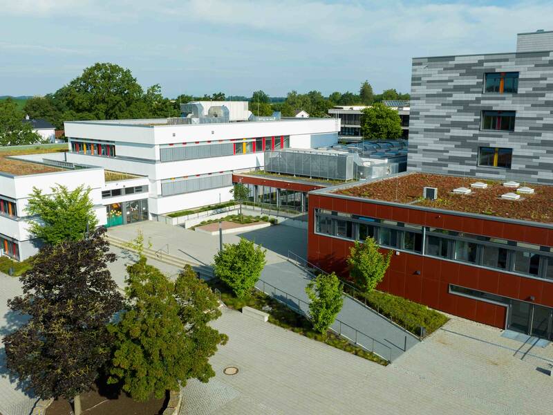 Ein moderner Gebäudekomplex mit Flachdachbegrünung und bunten Fenstern, umgeben von Bäumen und einer gepflasterten Fläche.