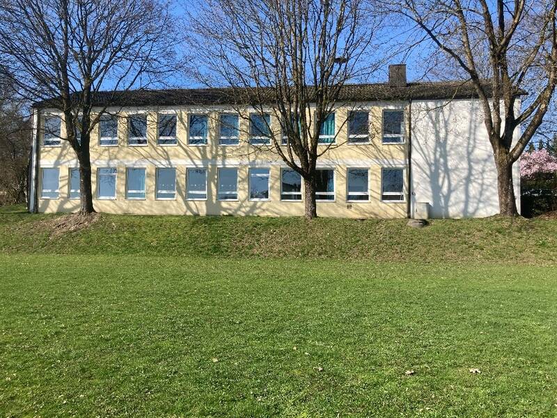Im Bild ist ein ein zweistöckiges Schulgebäude. Im Vordergrund sind mehrere Bäume.