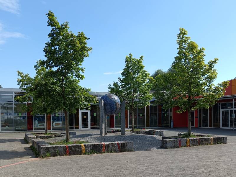 Vor einem Schulgebäude mit großen Fenstern und roten Wänden stehen mehrere Bäume, in deren Mitte eine Weltkugel aus Stein von Steinssulen gehalten.