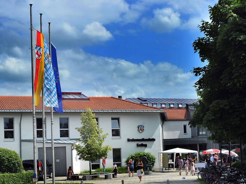 Ein zweistöckiges Gebäude mit roten Dachziegeln, davor drei Flaggenmasten und mehrere Personen sowie ein Baum und Fahrräder im Vordergrund.