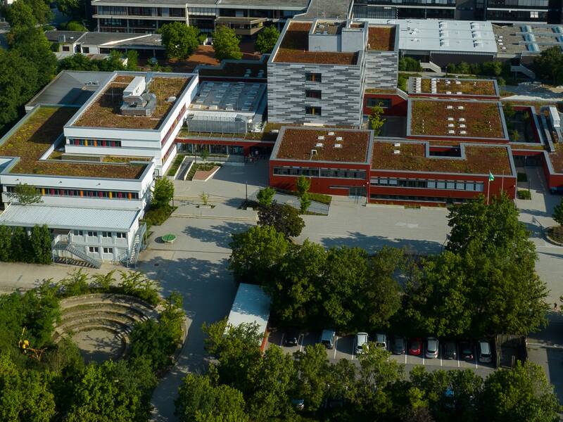 Ein moderner Gebäudekomplex mit Flachdachbegrünung und in rot grau, umgeben von Bäumen und einer gepflasterten Fläche.
