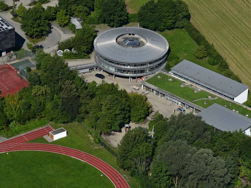 Luftaufnahme eines Schulkomplexes mit einem kreisförmigen Hauptgebäude, umgeben von Bäumen, Sportplätzen und einer Laufbahn.