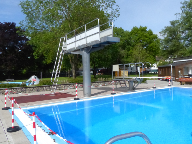 Ein Schwimmbad mit einem Sprungturm und einem Sprungbrett, umgeben von Bäumen und Gebäuden, abgesperrt durch rot-weiße Ketten.