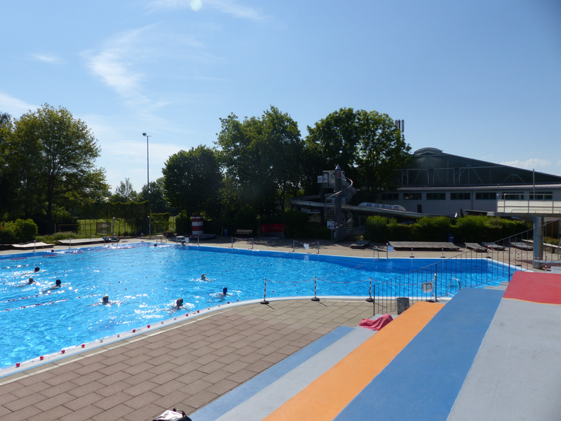 Ein Freibad bei sonnigem Wetter mit mehreren schwimmenden Personen. Dahinter eine Wasserrutsche.
