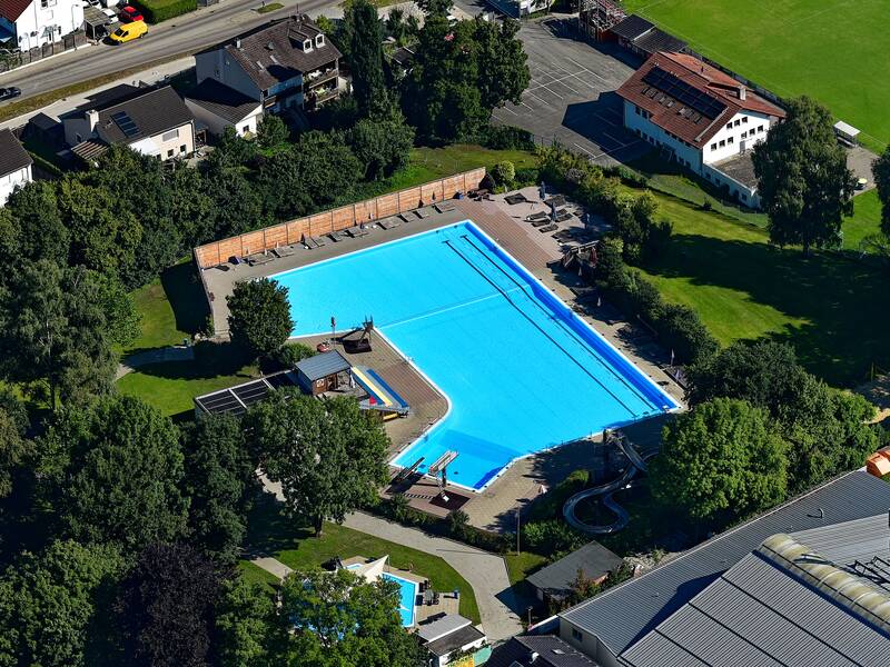 Eine Lufaufnahme von einem Freibad. Ein großes Becken in der Mitte, um gebend von grüner Wiese, Bäumen und einzelnen Häusern.n