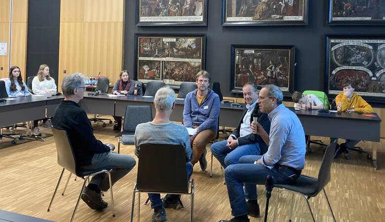 Ein Bild vom Jugend.Forum 2025 im Sitzungssaal des Rathauses der Stadt Dorfen. Es sitzen mehrere Menschen in einem Kreis in der Mitte, während andere an Tischen rundherum zuschauen.