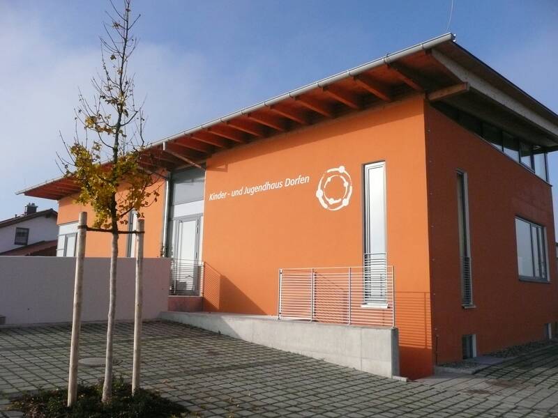 Ein modernes Gebäude mit oranger Fassade und der Aufschrift "Kinder- und Jugendhaus Dorfen" steht neben einem jungen Baum.