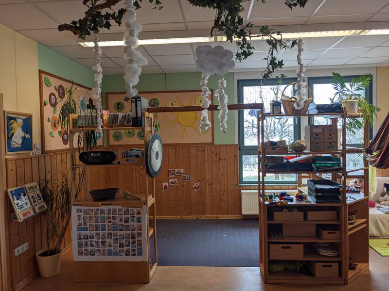 Ein bunter Kindergartenraum mit Holzregalen, dekoriert mit Wolken aus Watte und Pflanzen, bietet eine warme, einladende Atmosphäre mit vielen pädagogischen Materialien und gemalten Wandbildern.
