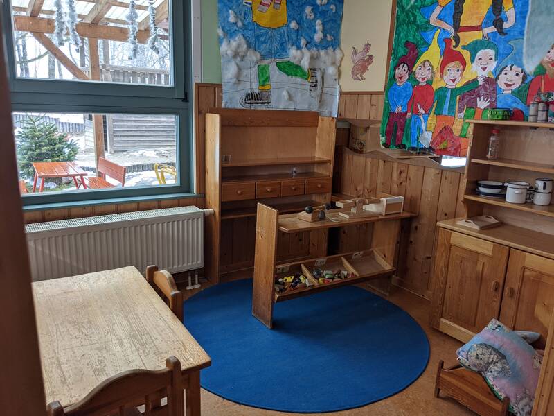 Ein Kindergartenraum mit Holzmöbeln, einem blauen Teppich und bunten Kinderzeichnungen an den Wänden; draußen steht ein Tisch unter einer überdachten Terrasse.