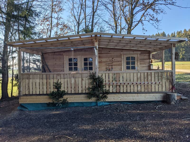 Eine hölzerne Hütte mit Veranda steht zwischen Bäumen auf einem ländlichen Grundstück.