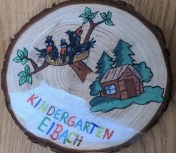 Eine bemalte Holzscheibe zeigt drei schwarze Vögel in einem Nest auf einem Baum sowie eine kleine Hütte und Kiefern, mit der Aufschrift "KINDERGARTEN EIBACH" in bunten Buchstaben.