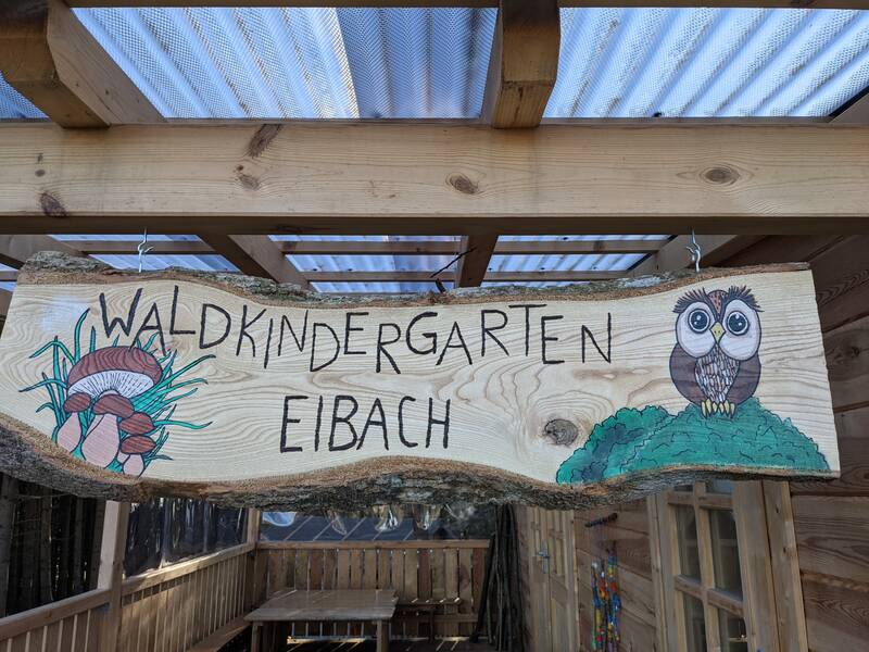 Ein Holzschild mit der Aufschrift "Waldkindergarten Eibach" zeigt Zeichnungen von Pilzen auf der linken Seite und einer Eule auf der rechten Seite.
