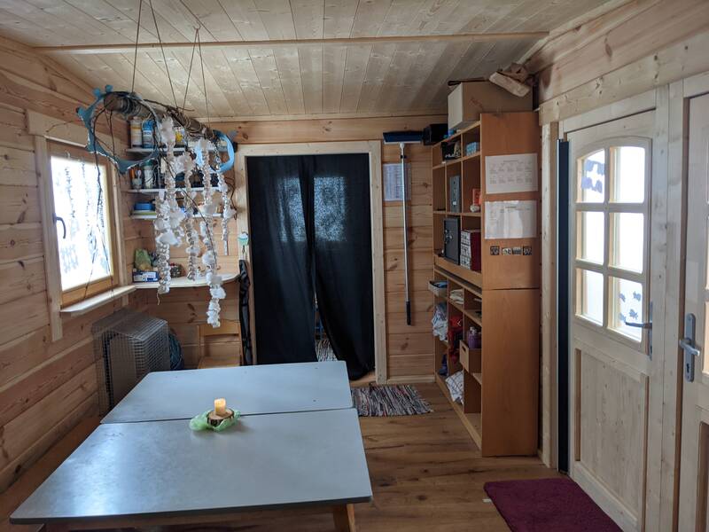 In einem kleinen, holzverkleideten Raum stehen ein Tisch mit einer brennenden Kerze, einige Regale voller Gegenstände und eine Tür mit einem Fenster, das teilweise abgedeckt ist.