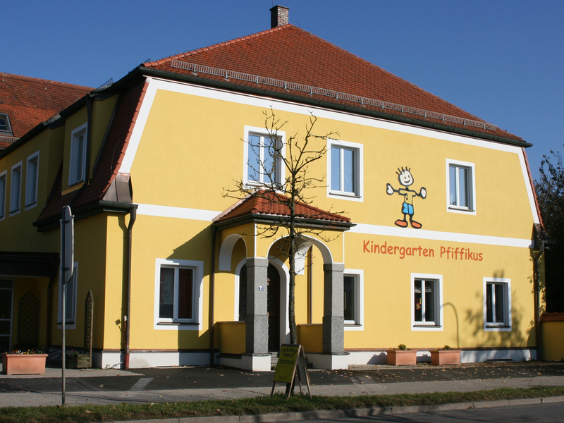 Ein gelbes Gebäude mit rotem Dach, auf dem "Kindergarten Pfiffikus" steht, ist mit einer bunten Zeichnung eines fröhlichen Kindes dekoriert.