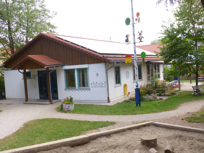 Ein Gebäude mit einem roten Ziegeldach und bemalten Figuren auf einem Maibaum steht in einer grünen, gepflegten Umgebung mit einem Sandkasten im Vordergrund.