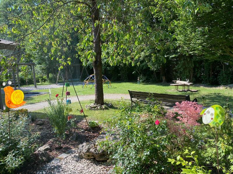 Ein grüner Garten mit Bäumen, bunten Glasmosaiken, einer Bank und Spielgeräten.