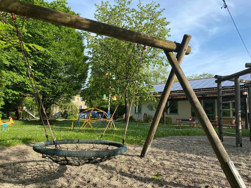 Ein Spielplatz mit einer Nestschaukel, Klettermöglichkeiten und einem Gebäude im Hintergrund bei sonnigem Wetter.