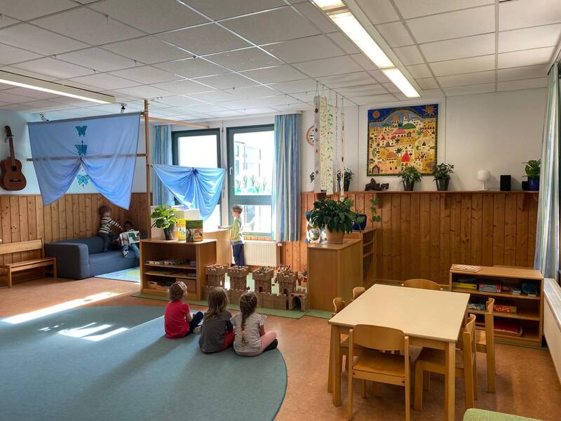 Ein heller Kindergartenraum mit Holztischen und -stühlen, einem Teppich, mehreren Pflanzen, Spielzeug und Kindern, die spielen.