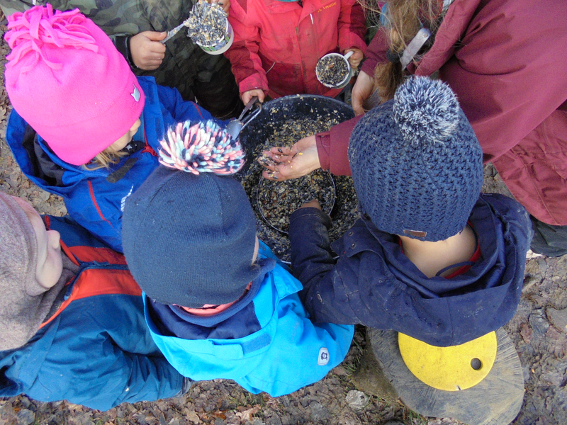 Mehrere Kinder in bunten Winterjacken und Mützen sind im Freien versammelt und mischen etwas in einer großen Schüssel.