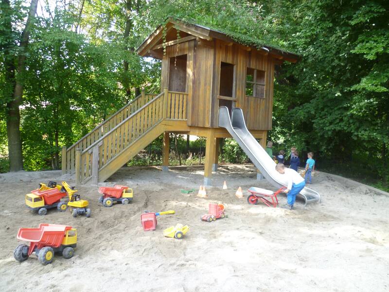 Auf einem Spielplatz mit Sandkasten steht ein Holzspielhaus mit Rutsche, umgeben von Spielzeugfahrzeugen, während vier Kinder spielen.