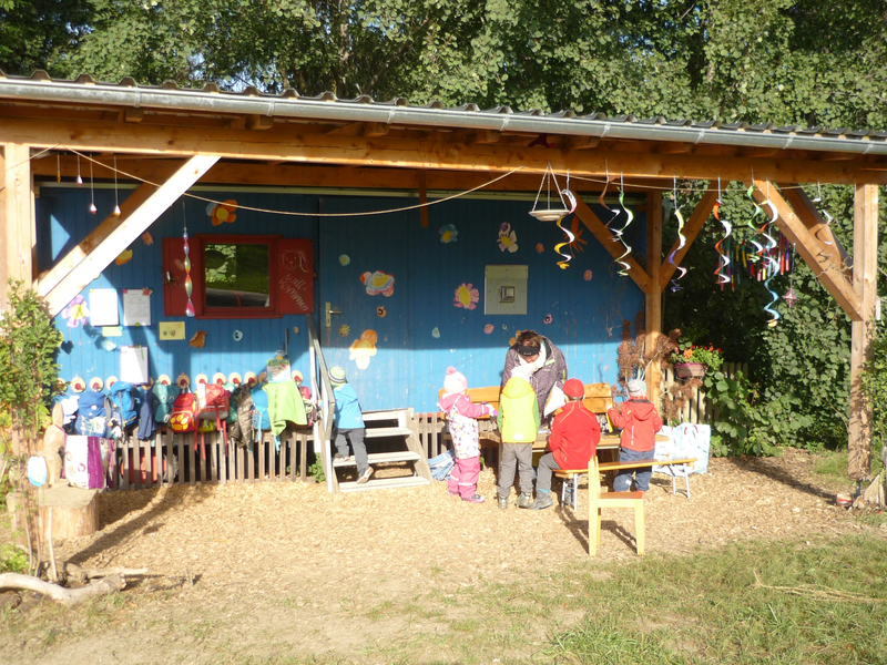 Eine Gruppe von Kindern steht mit einer erwachsenen Person vor einer bunt dekorierten blauen Holzhütte im Freien.