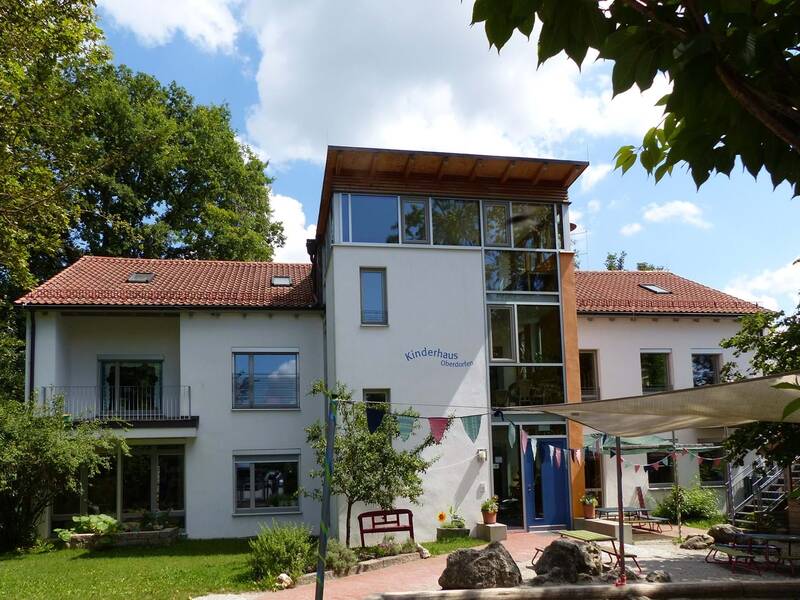 Ein großes zweistöckiges Haus mit einem Vorbau in der Mitte auf dem "Kinderhaus Oberdorfen" steht. Vor dem Haus sind ein Sonnensegel und Wimpel die den Picknickplatz einrahmen.