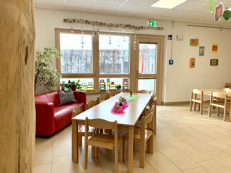 Ein Raum mit einem großen Tisch mit Stühlen eine roten Couch links und bunten Wanddekorationen neben großen Fenstern.