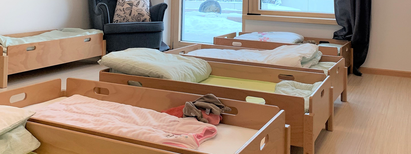 Ein heller Raum mit mehreren niedrigen Kinderbetten aus Holz, einem Sessel in der Ecke und einem großen Fenster im Hintergrund.