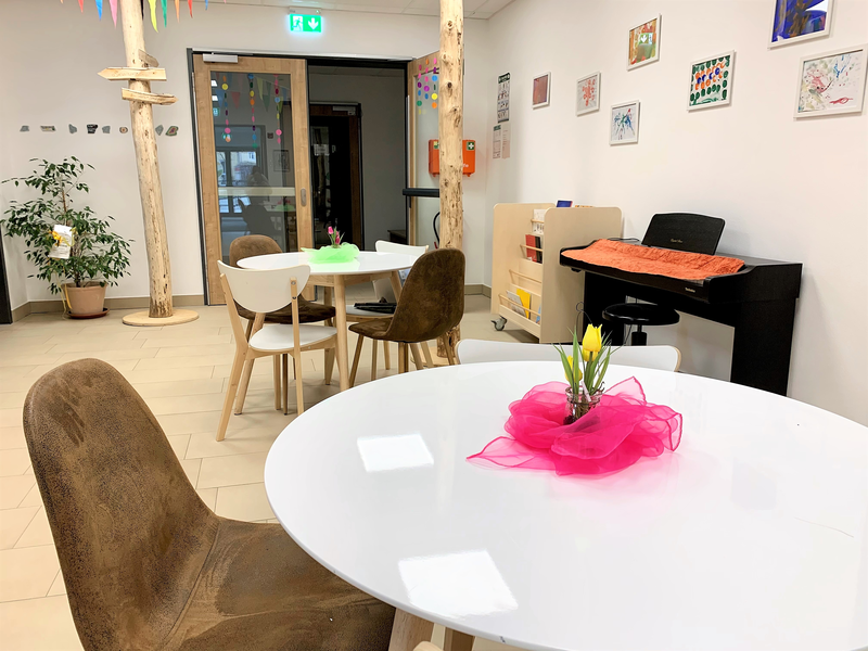 Ein heller Raum mit weißer Wand ist möbliert mit runden Tischen, bunten Blumenarrangements und verschiedenen Stühlen; im Hintergrund ist ein Piano und an der Wand hängen bunte Bilder.