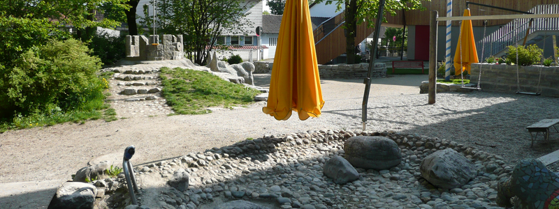 Ein Spielplatz mit großen Steinen, zwei gelben Sonnenschirmen einem Maibaum und grünen Bäumen im Hintergrund.