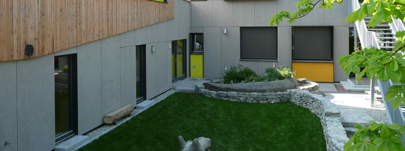 Ein modernes Gebäude mit einer Fassade aus Holz und grauen Paneelen, umgeben von einer Grünfläche und einem gepflegten Garten.