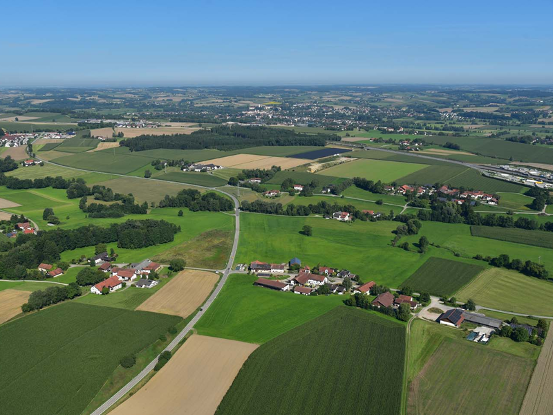 Luftaufnahme einer ländlichen Landschaft mit weitläufigen Feldern, verstreuten Wohnhäusern und kleinen Wäldern unter einem klaren blauen Himmel.
