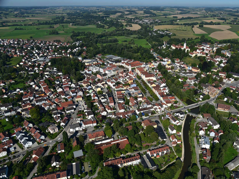 Luftaufnahme der Stadt Dorfen mit vielen dicht beieinander liegenden Gebäuden, einem Fluss und umgeben von weitläufigen landwirtschaftlichen Flächen.