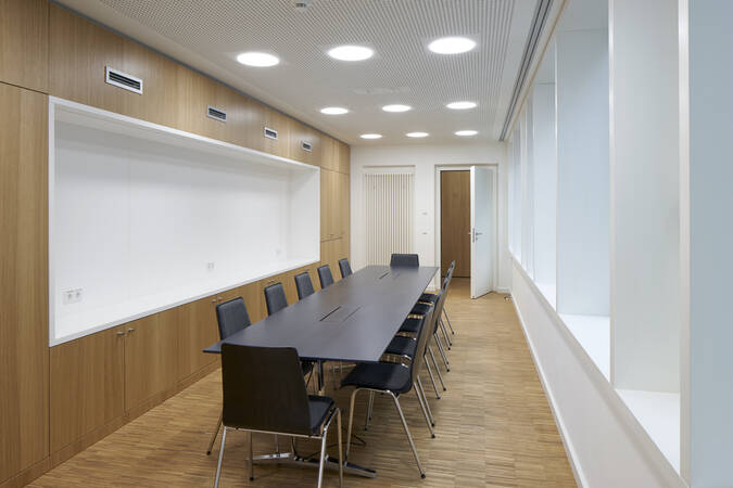 Ein moderner Konferenzraum mit einem langen Tisch und mehreren schwarzen Stühlen, umgeben von Holzverkleidungen und großen Fenstern auf der rechten Seite.