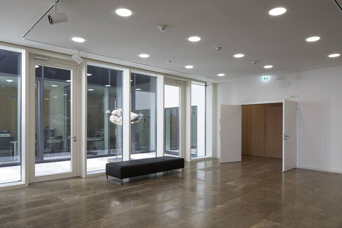 Ein heller Raum mit großen Glasfenstern, einer schwarzen Bank in der Mitte und einer offenen Tür auf der rechten Seite.