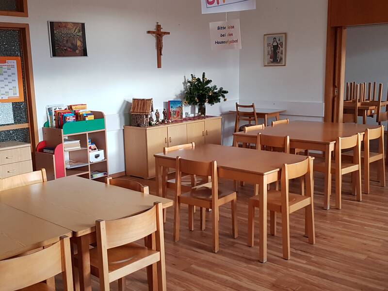 Ein heller Raum mit mehreren Holztischen und Stühlen, einem Schrank voller Bücher und Spielzeug, sowie religiösen Symbolen an den Wänden.