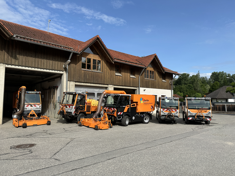 Fünf orangefarbene Servicefahrzeuge mit verschiedenen Geräten und Aufbauten stehen vor einem großen Gebäude mit hölzernen Fassaden und roten Dächern.