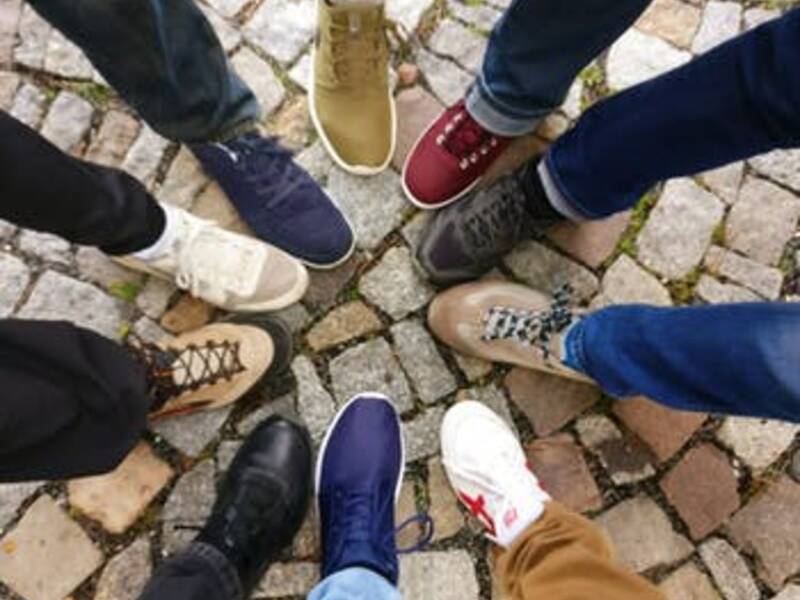 Eine Gruppe von acht Menschen steht im Kreis auf einem Kopfsteinpflaster, wobei nur ihre verschiedenfarbigen Schuhe sichtbar sind.