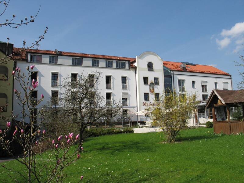Großes, weißes vier-stöckiges Gebäude. Vornedran ein Garten mit Bäumen und Sträuchern.
