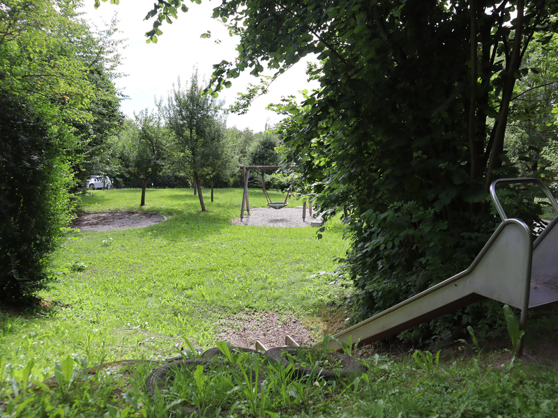 Ein grüner Spielplatz mit einer Rutsche im Vordergrund und Schaukeln im Hintergrund, umgeben von Bäumen.