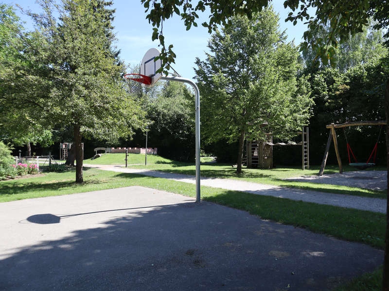 Ein sonniger Spielplatz mit einem Basketballkorb, einer Schaukel und einem Spielturm, umgeben von vielen Bäumen.